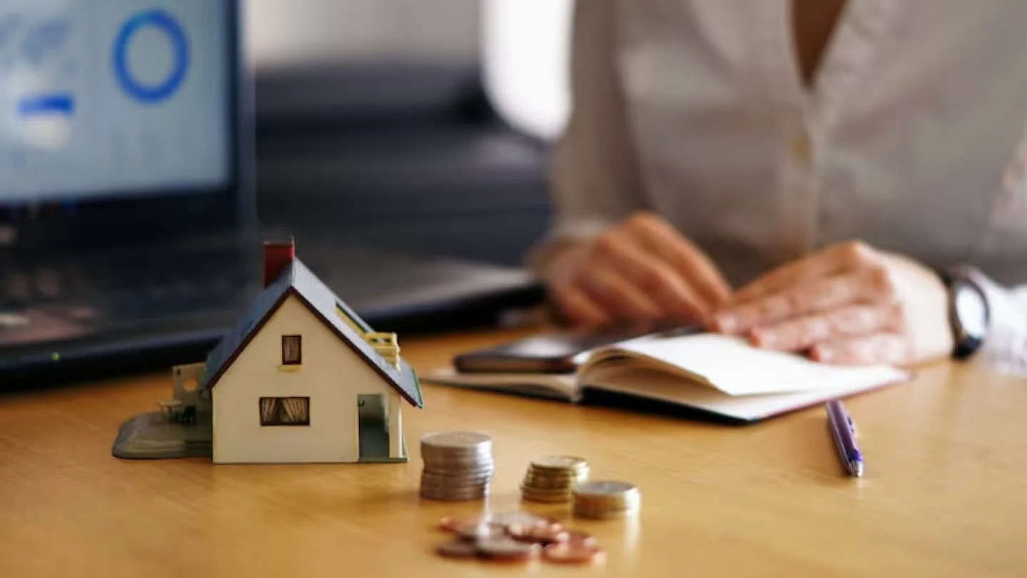 Modelo de casa junto a monedas y una laptop en un escritorio