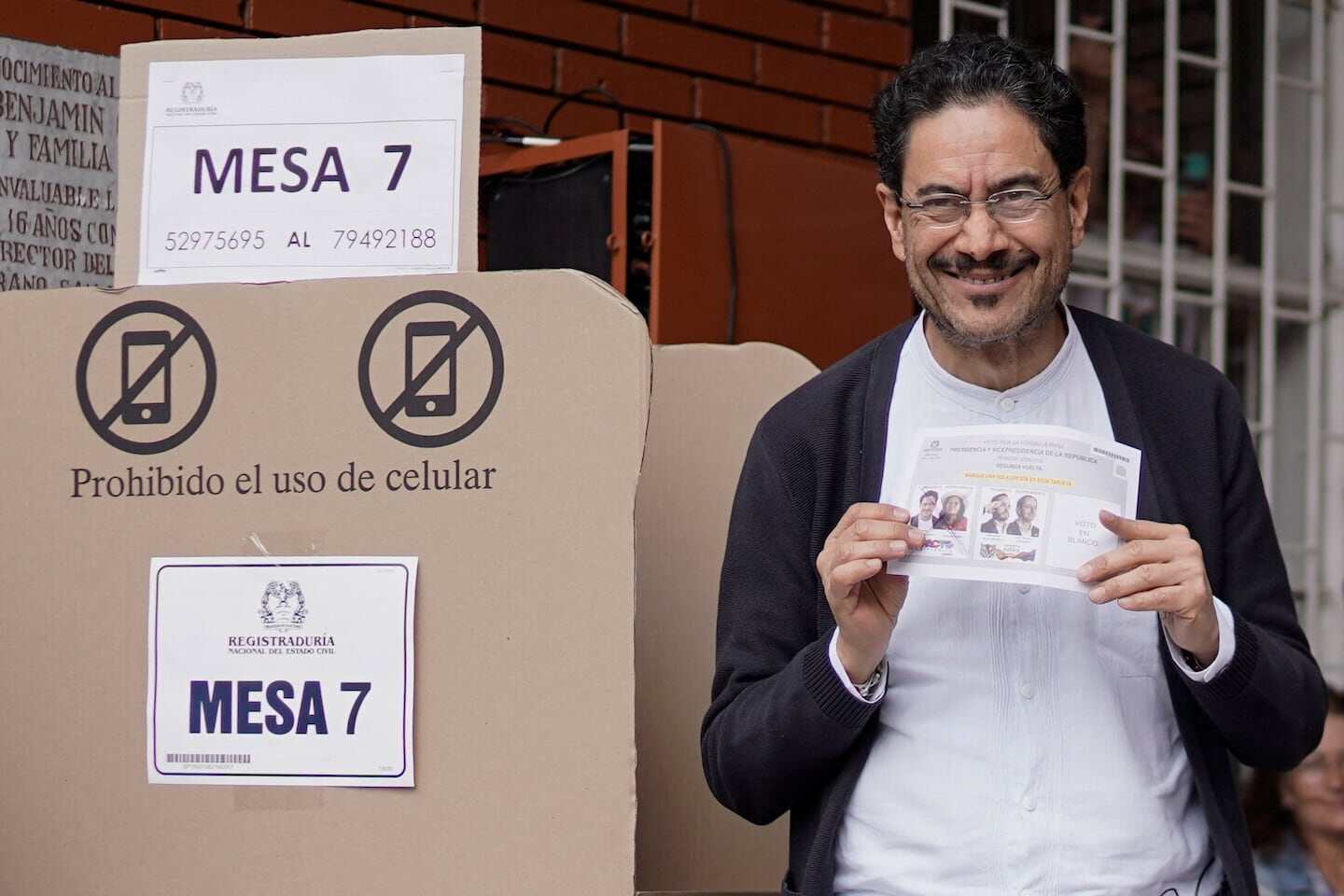 Hombre sonriente sosteniendo una boleta electoral frente a una mesa de votación en Colombia