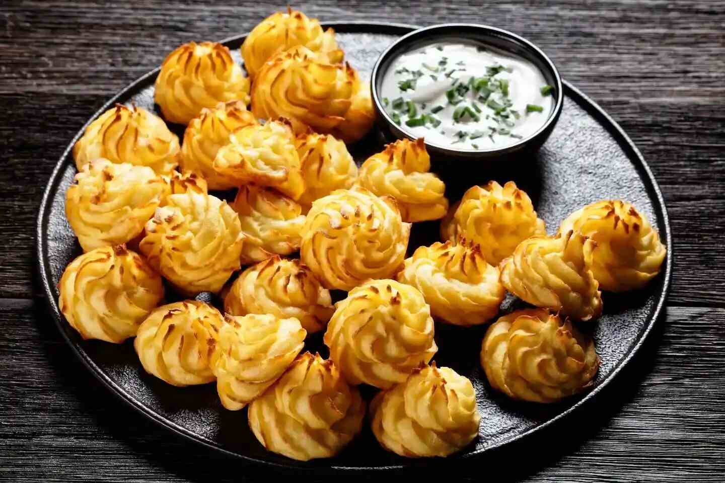 Papas duquesas doradas en un plato negro con salsa de acompañamiento