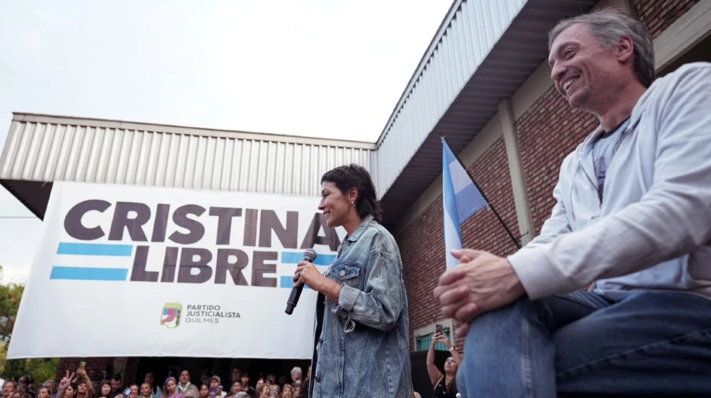 Mujer hablando en un mitin político con un hombre sentado a su lado y un gran cartel que dice 'Cristina Libre' en el fondo
