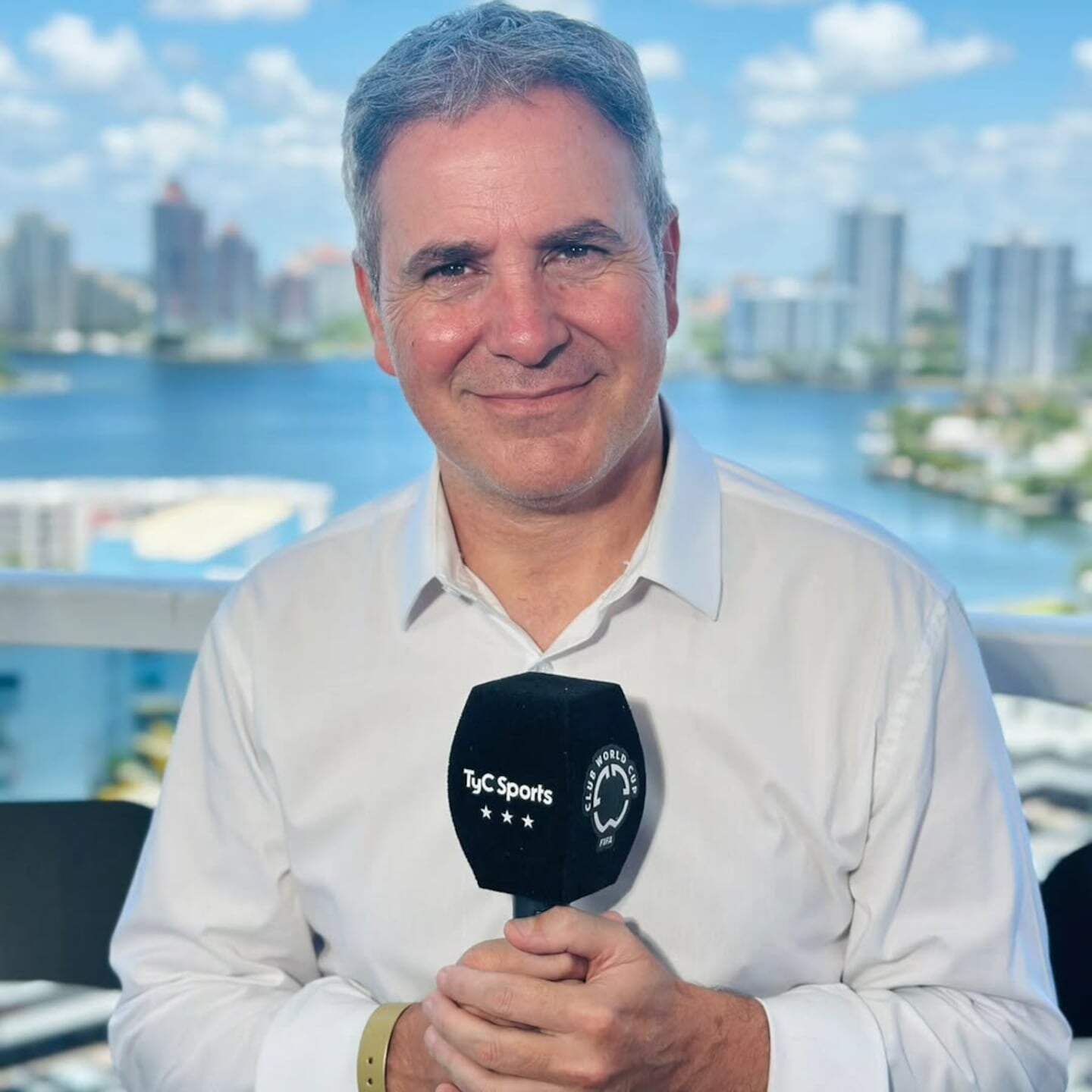 Ariel Rodríguez sonríe mientras sostiene un micrófono de TyC Sports frente a un paisaje urbano