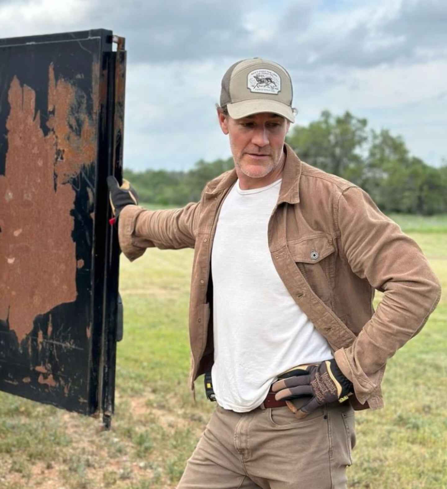 James Van Der Beek en un entorno rural, vestido con chaqueta y gorra, apoyado en una puerta metálica