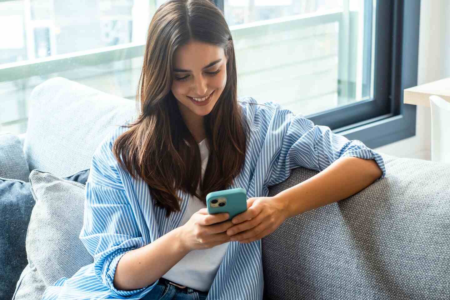Joven sonriendo mientras usa su celular en un sofá