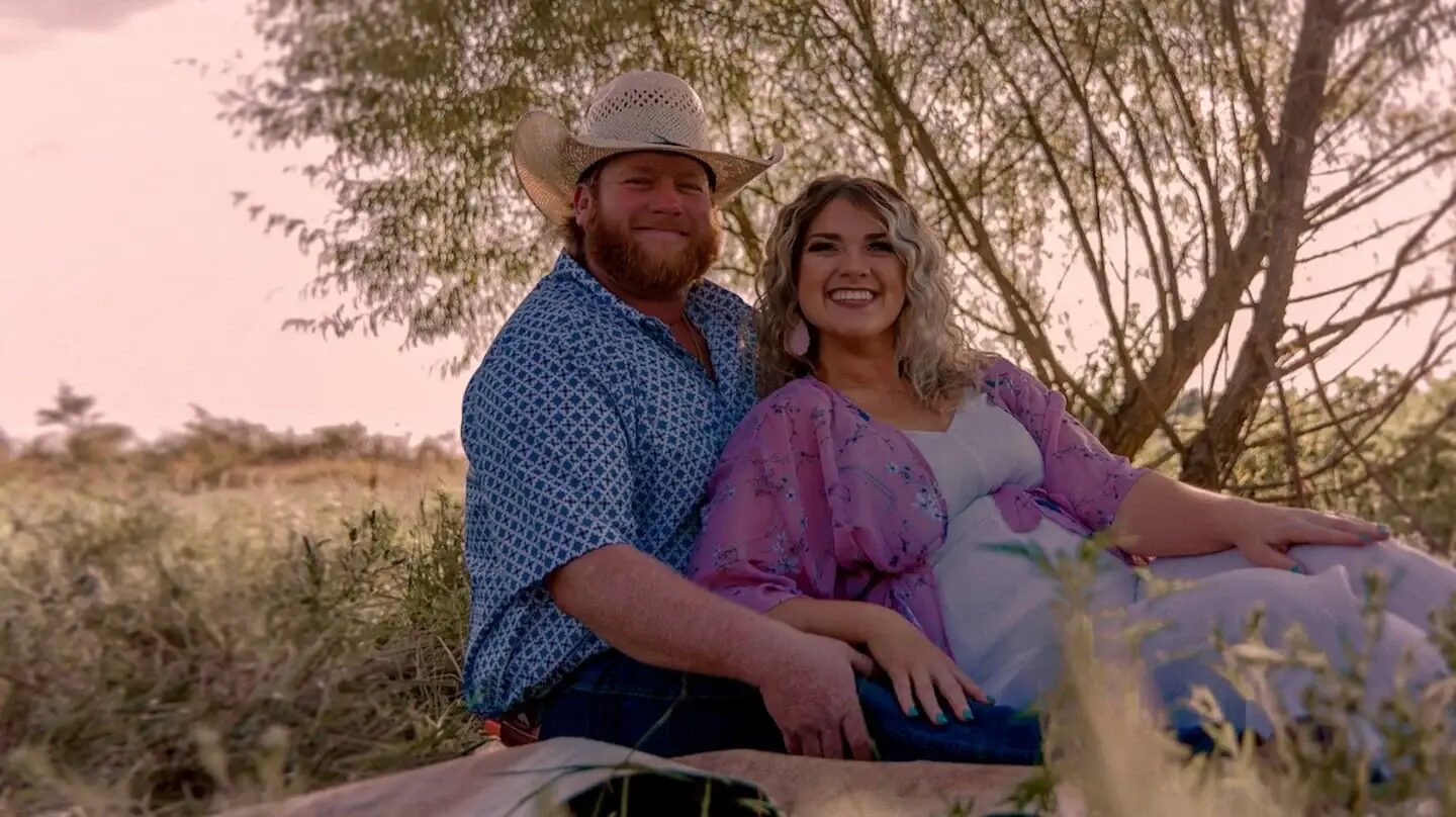 Pareja sonriente en un entorno natural, con la mujer embarazada sentada junto a su pareja