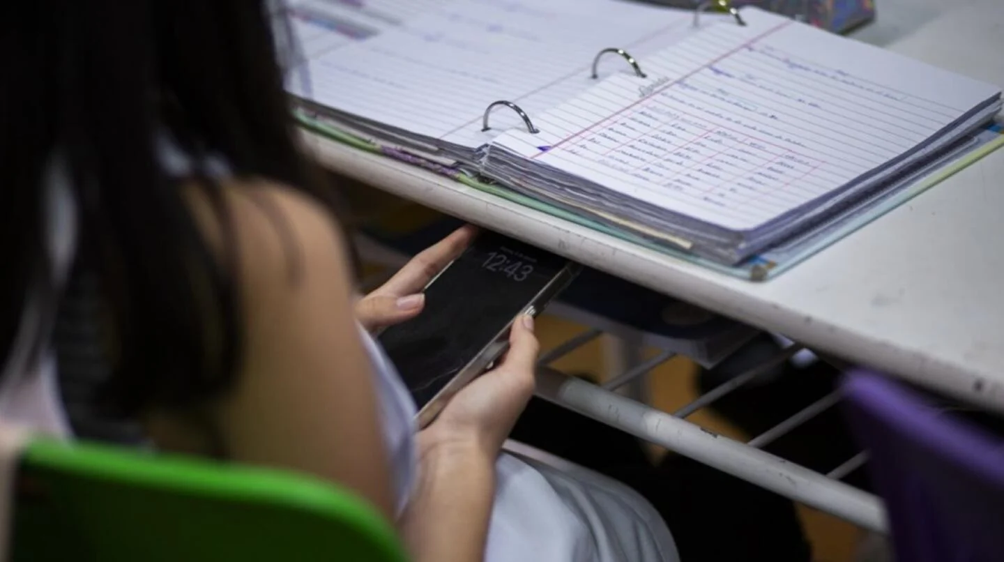 Estudiante sosteniendo un celular en un aula con cuadernos y carpetas en la mesa