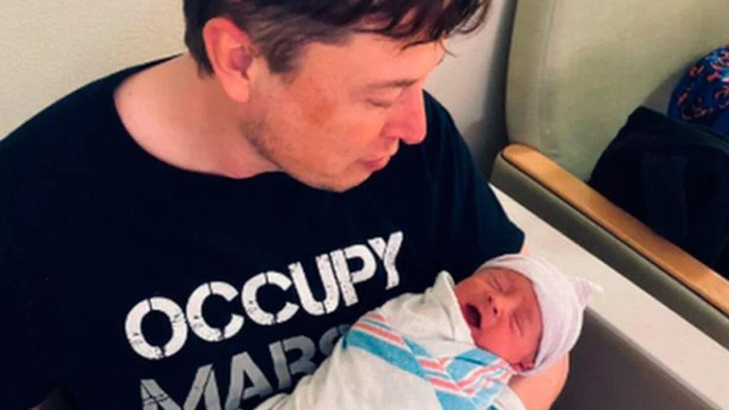 Elon Musk sosteniendo a un bebé en brazos mientras viste una camiseta con el lema 'OCCUPY MARS'