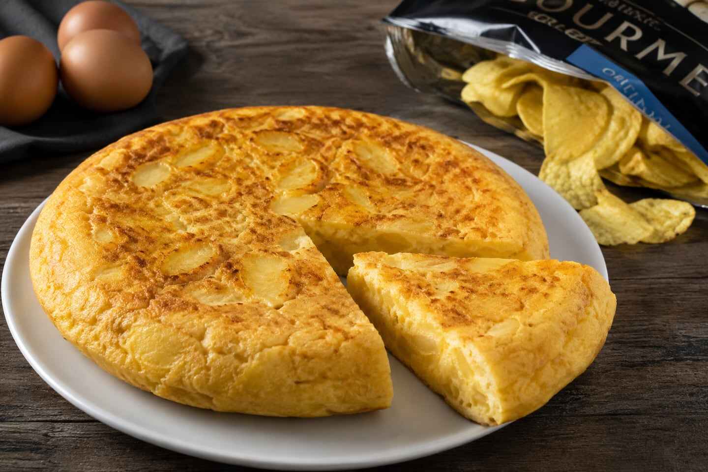 Tortilla de papas presentada en un plato con una porción cortada y papas fritas de paquete al fondo