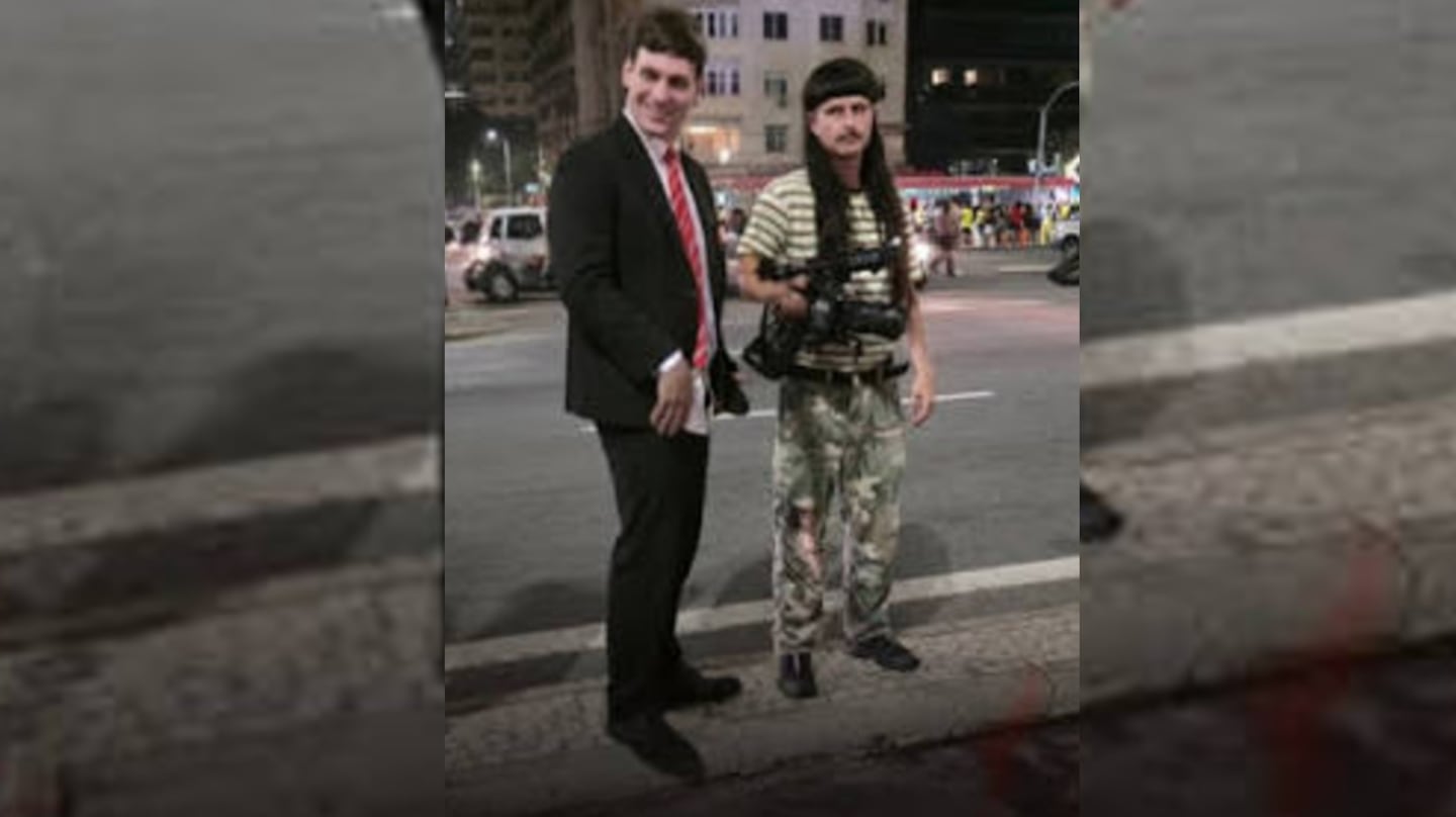Dos hombres posando en una calle de Río de Janeiro, uno vestido de traje y otro con ropa camuflada y una cámara