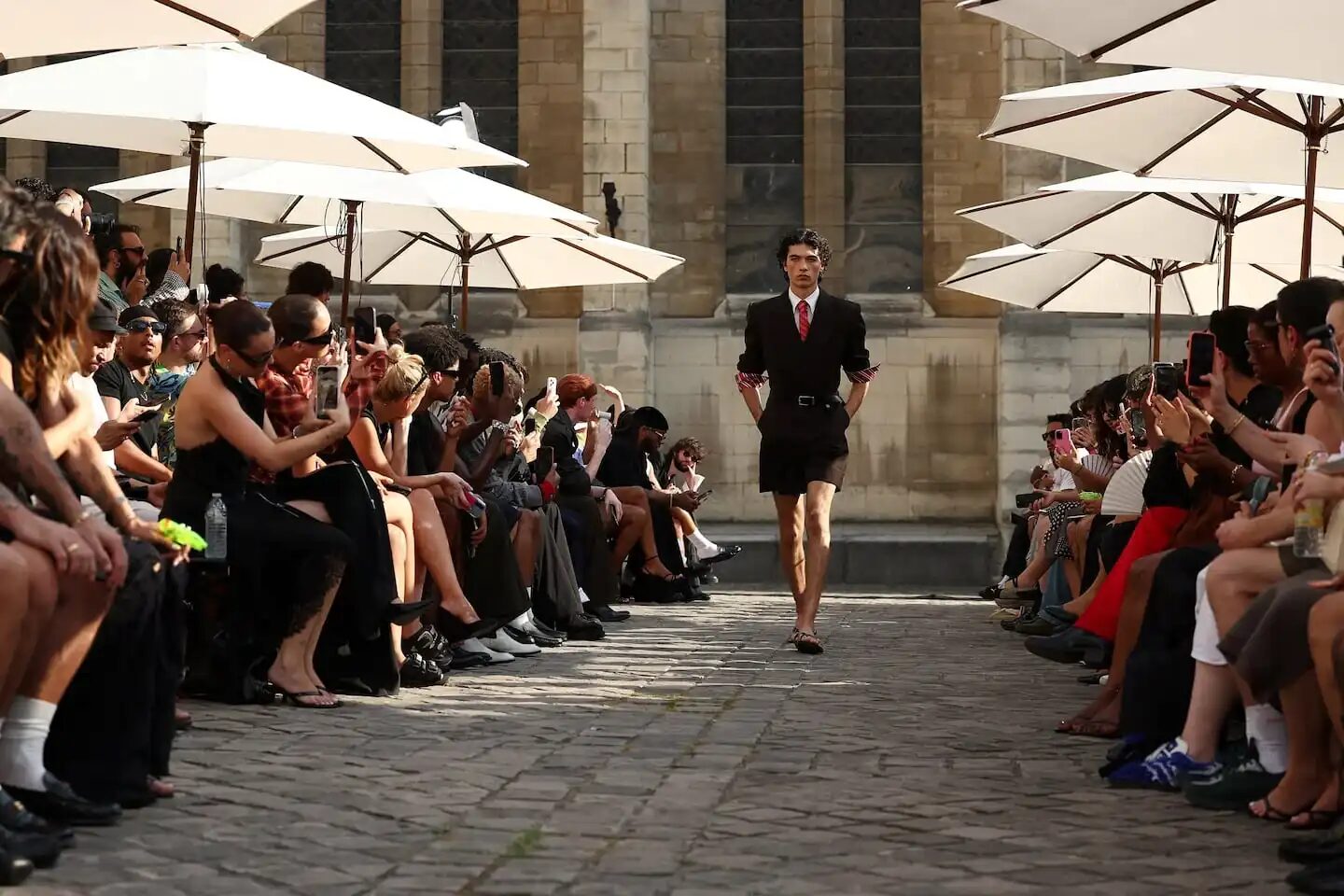 Modelo desfilando en la Semana de la Moda Masculina de París bajo sombrillas en un ambiente caluroso
