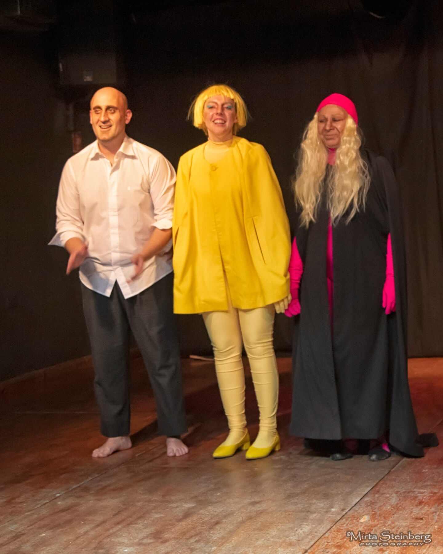 Tres actores en el escenario de la obra 'Querencio', vestidos con trajes coloridos y expresiones alegres