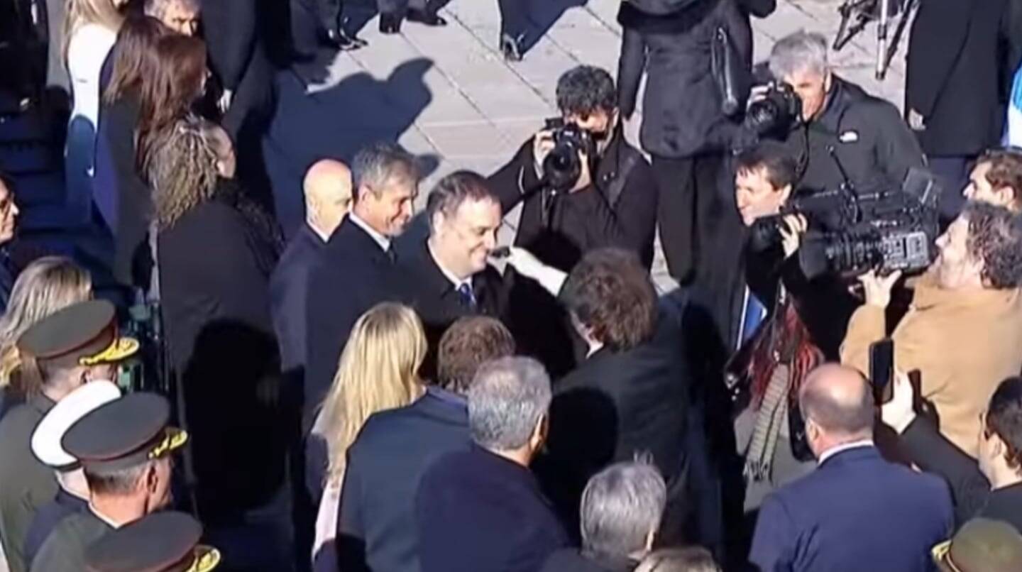 Manuel Adorni saluda a Javier Milei en un evento público rodeado de periodistas y funcionarios