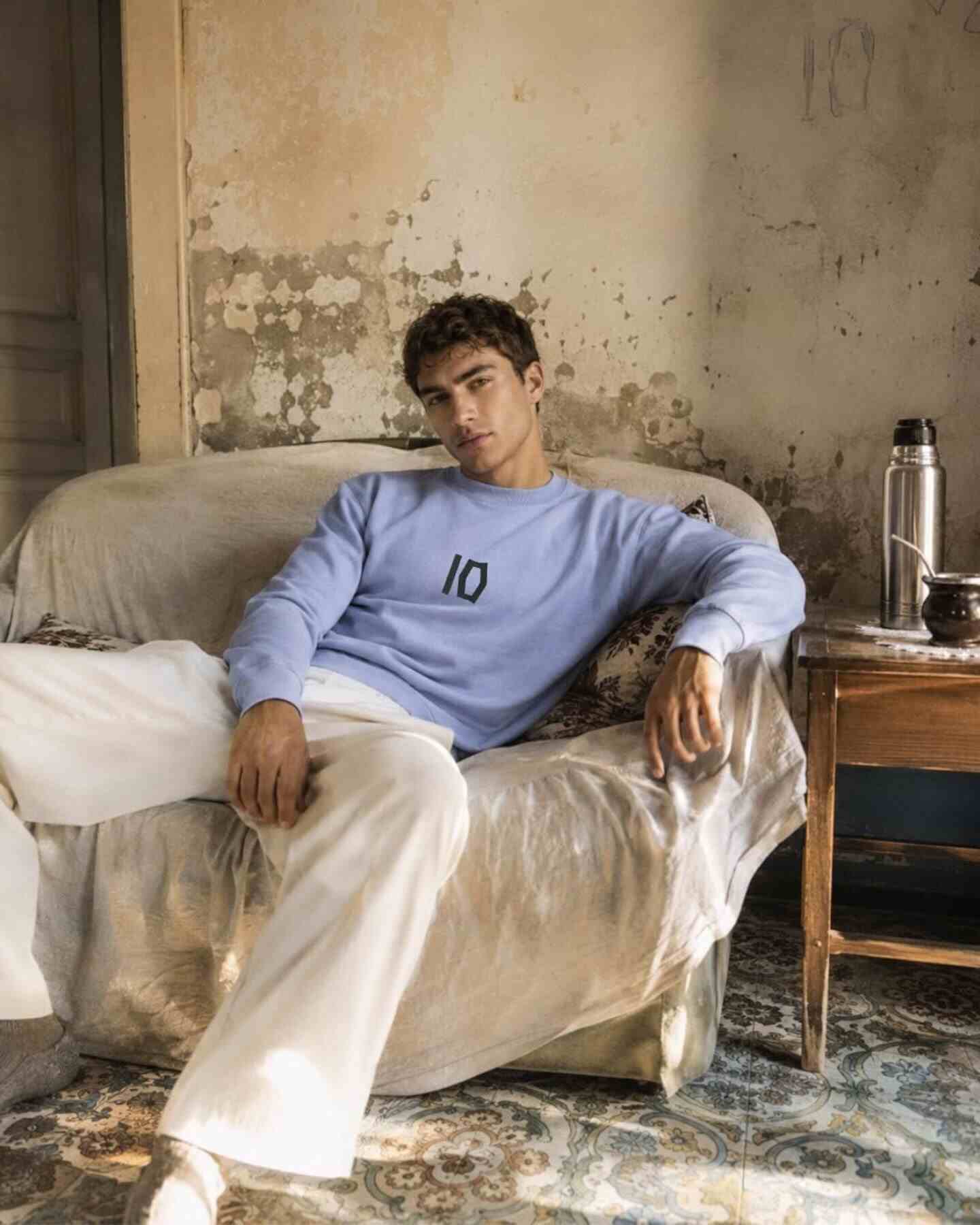 Modelo sentado en un sofá con sudadera azul y pantalones claros, mostrando el número 10 en la prenda