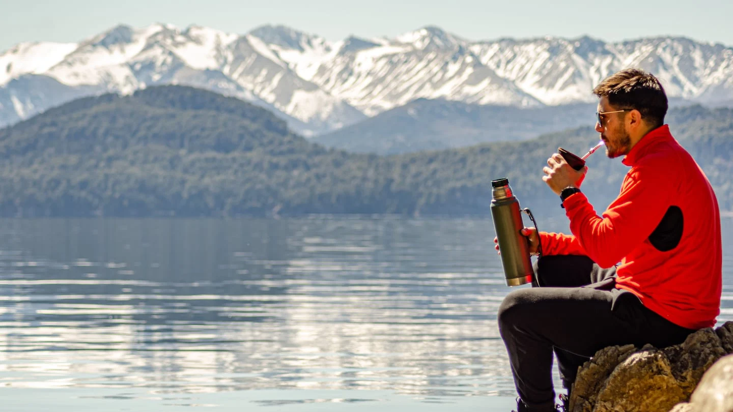 Hombre disfrutando de una bebida en la orilla de un lago con montañas nevadas al fondo