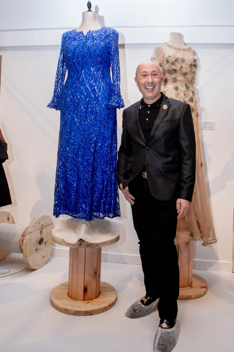 Claudio Cosano posando junto a un vestido azul brillante en una exhibición de moda