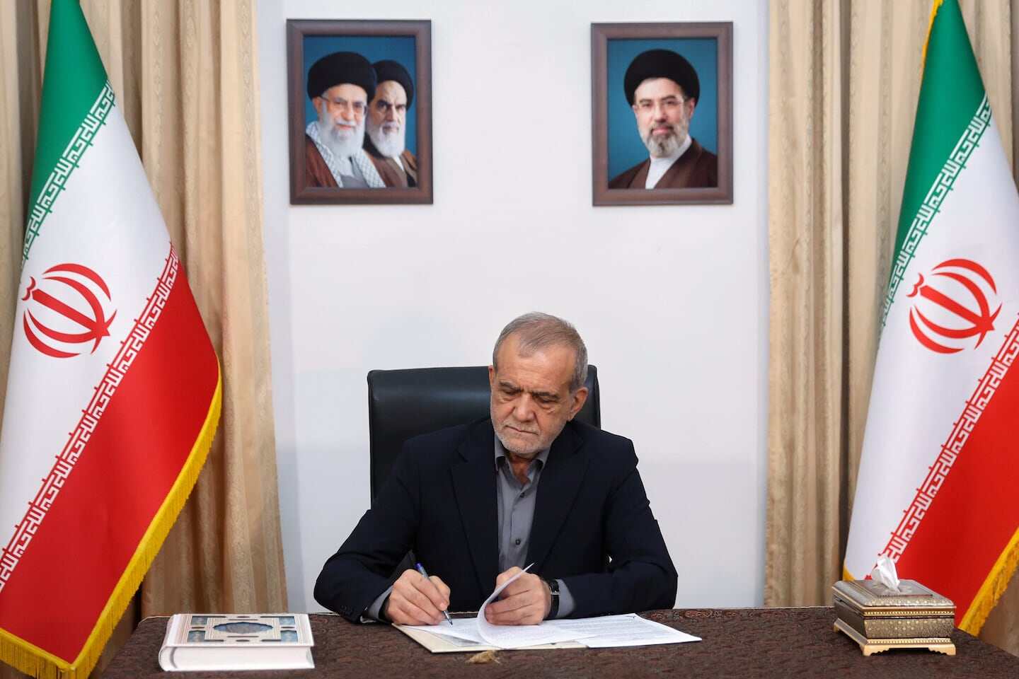 Rafael Grossi firmando documentos en una oficina con banderas de Irán de fondo