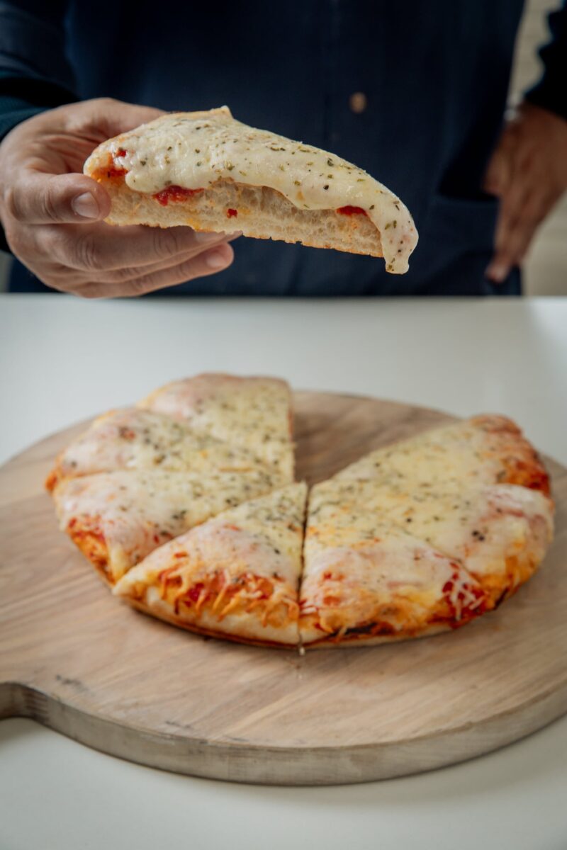 Pizza al molde lista para hornear sostenida por una mano, con una porción cortada en una tabla de madera