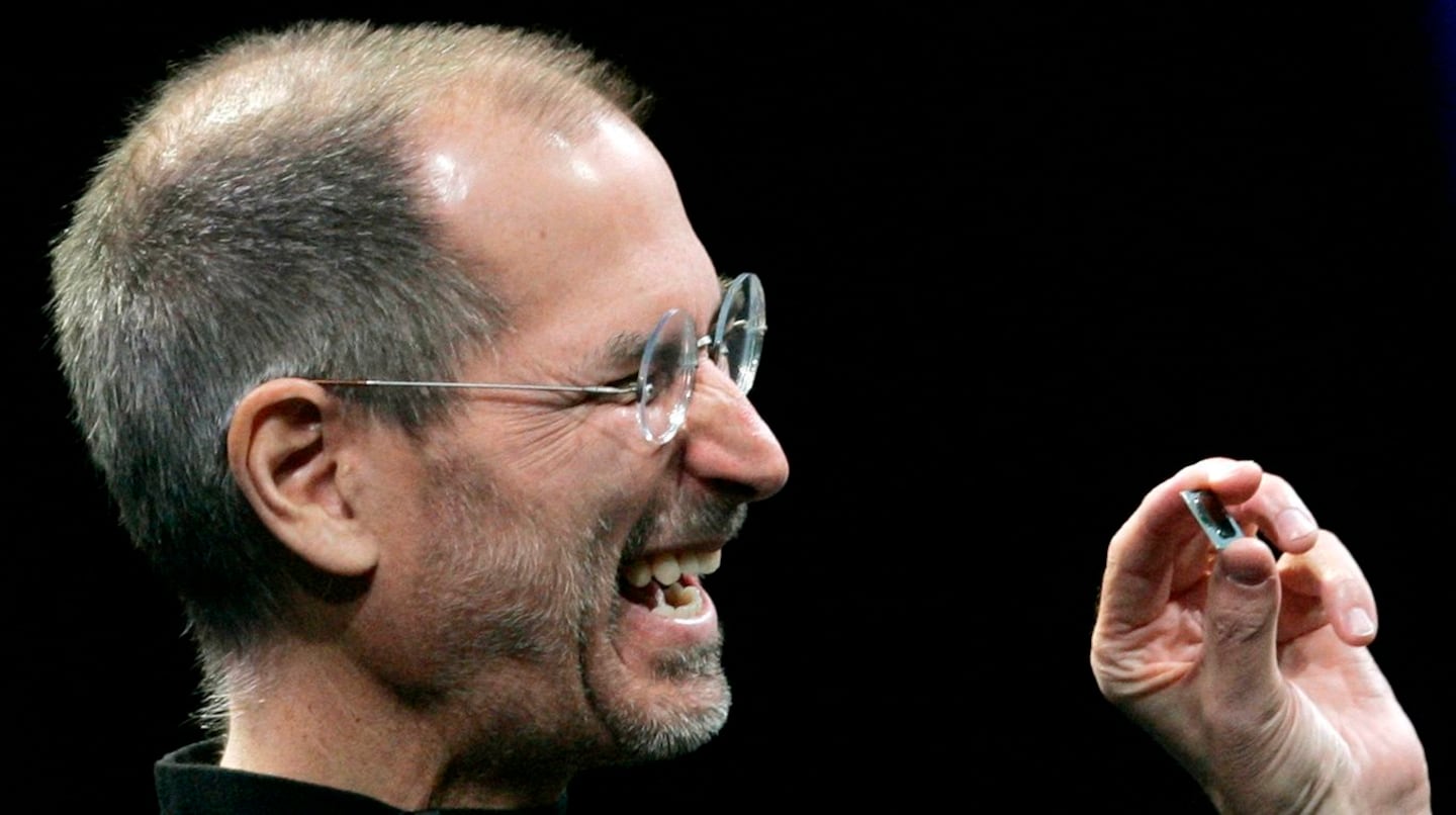 Steve Jobs sonriendo mientras sostiene un pequeño objeto en su mano durante una presentación