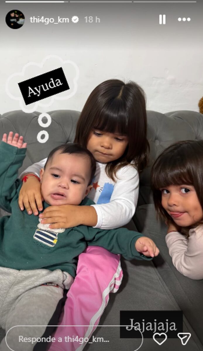 Tres niños posando en un sofá, uno de ellos sostiene a un bebé mientras los otros dos miran hacia la cámara