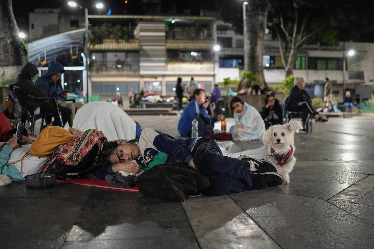 Hombre durmiendo en el suelo de una plaza con su perro, rodeado de personas en situación de emergencia tras un terremoto en Venezuela