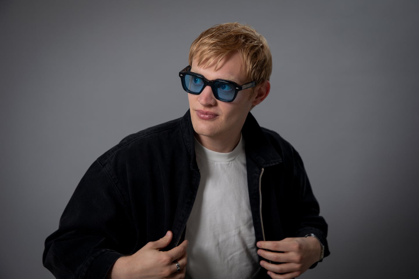 Joven con gafas de sol y chaqueta negra posando frente a un fondo gris