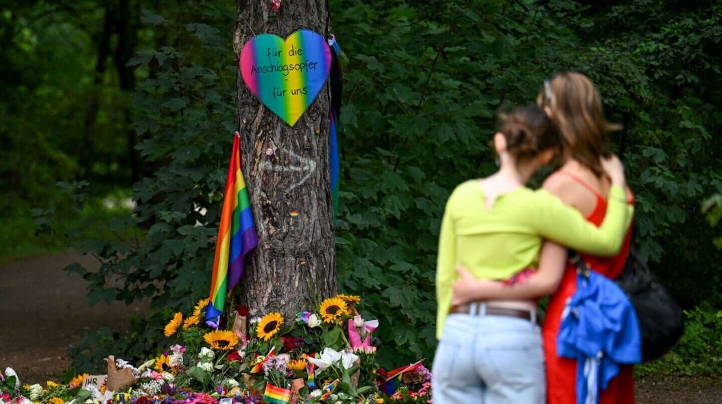 Dos personas se abrazan frente a un memorial con flores y banderas del Orgullo en Berlín
