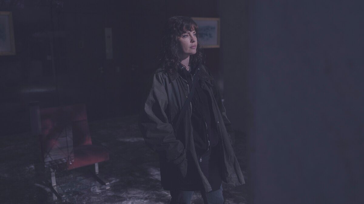 Mujer de pie en un ambiente oscuro y desolado, vestida con abrigo y mirada pensativa