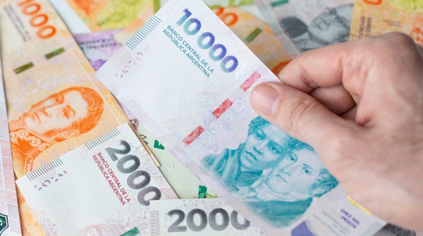 Una mano sostiene un billete de 10,000 pesos argentinos sobre una mesa con otros billetes de diferentes denominaciones