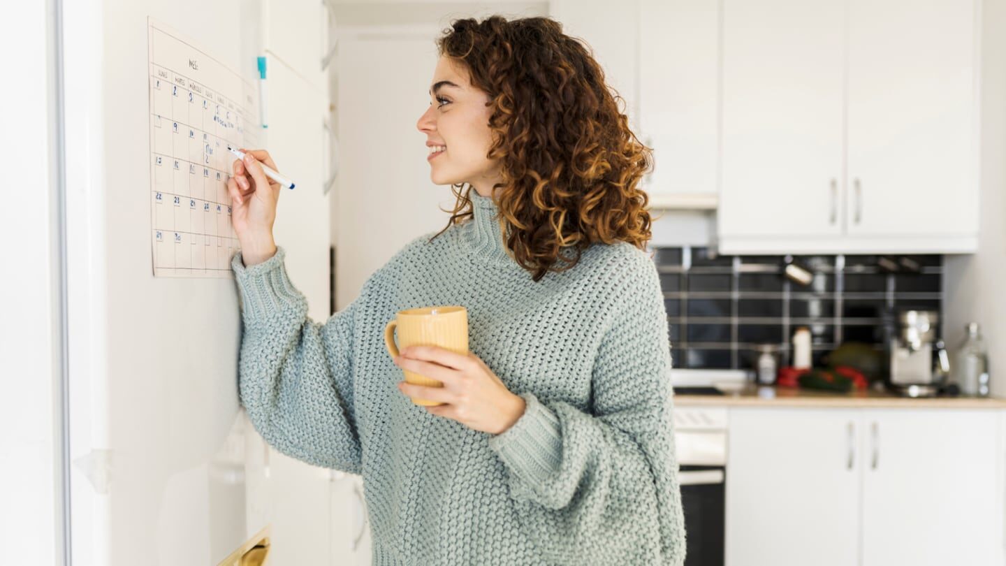 Mujer escribiendo en un calendario de papel mientras sostiene una taza en la mano