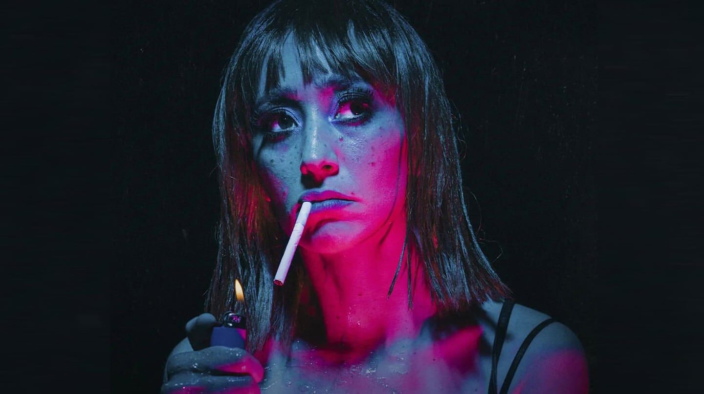 Mujer con cabello largo y lacio sosteniendo un cigarrillo y un encendedor en un ambiente oscuro y colorido