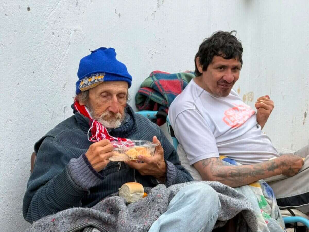 Dos hombres sentados en el exterior de un geriátrico clandestino en Santa Fe, uno comiendo y el otro mirando a la cámara
