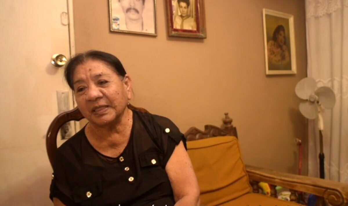 Mujer mayor sentada en un sofá con fotografías en la pared detrás de ella