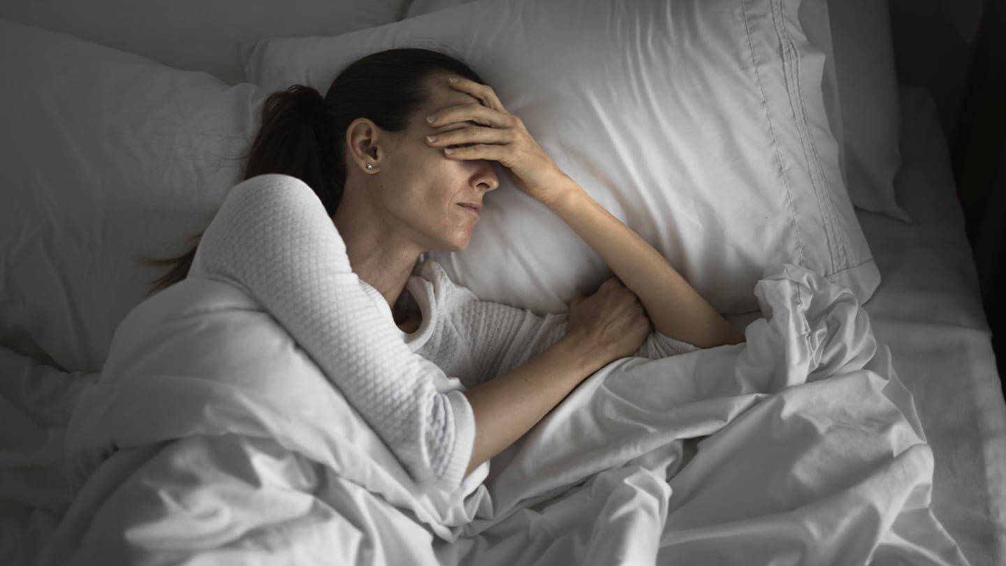 Mujer con expresión de angustia en la cama, cubriéndose con sábanas blancas