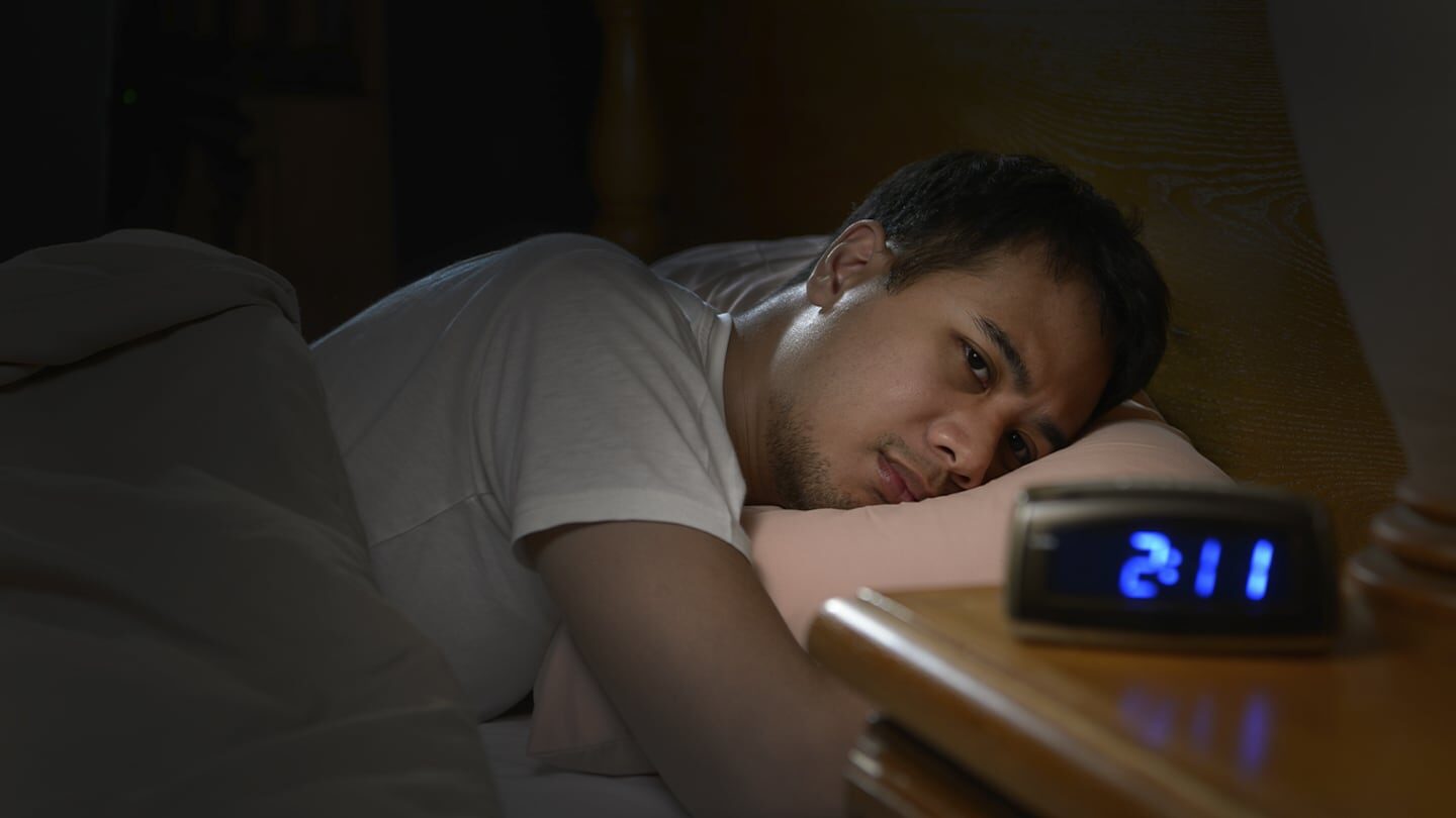 Hombre desvelado mirando un reloj despertador en la cama