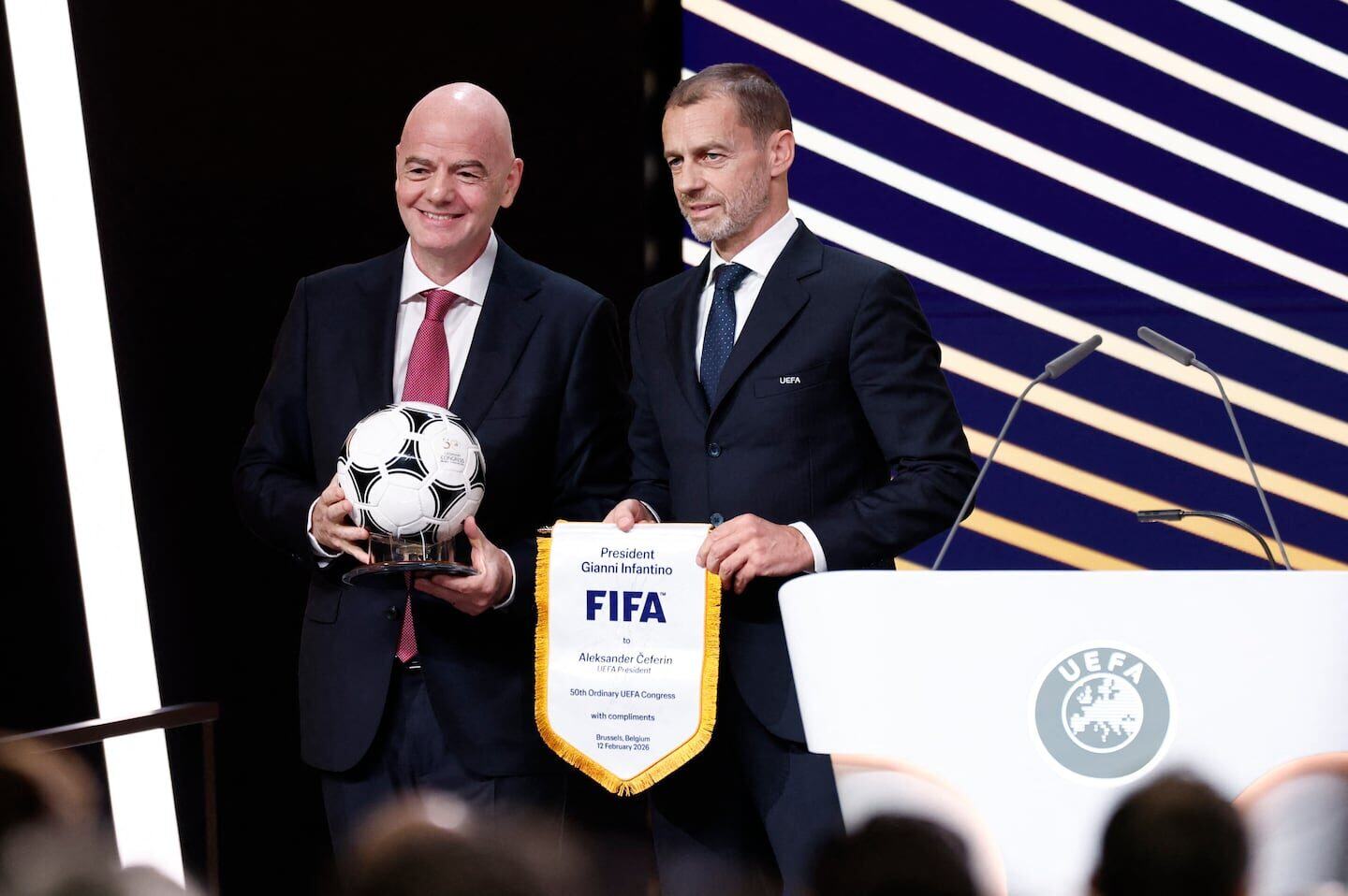 Gianni Infantino y Aleksander Čeferin en un evento de la UEFA sosteniendo un balón de fútbol y un estandarte