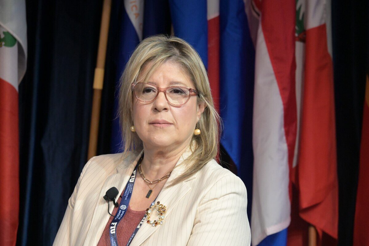 Mujer con gafas y chaqueta clara, en un evento con banderas de fondo