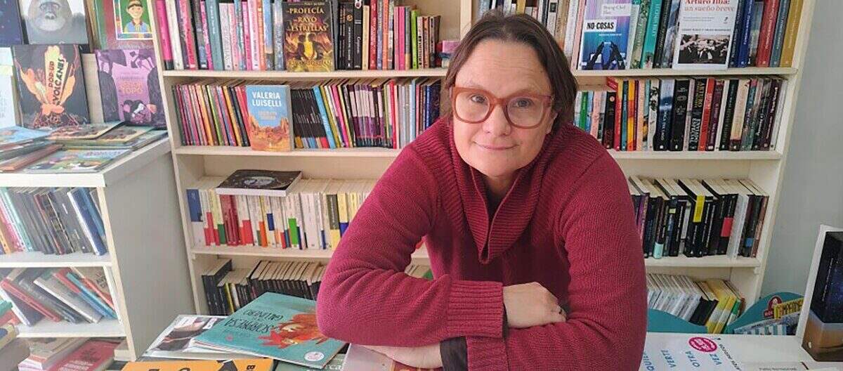Mujer con gafas y suéter rojo sonríe en una librería con estanterías llenas de libros