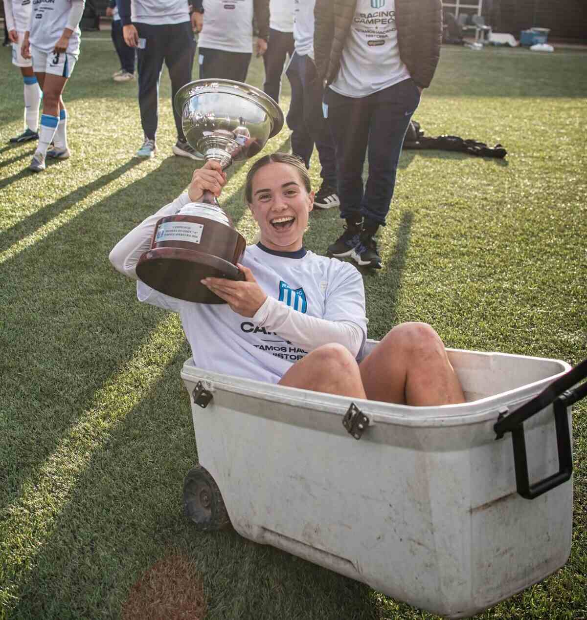 Jugadora de Racing Club sosteniendo el trofeo del Torneo Apertura mientras está sentada en un recipiente blanco