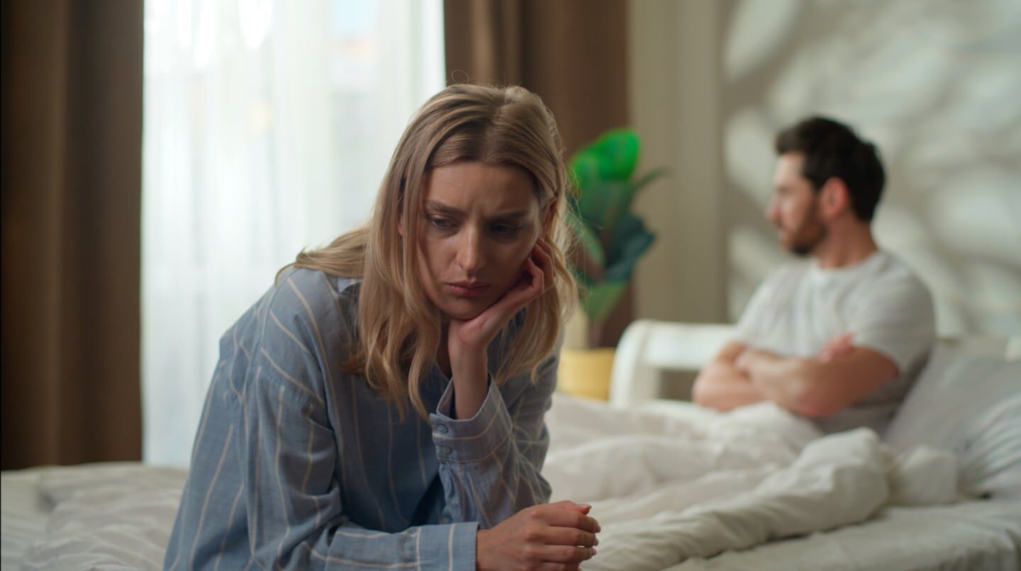 Pareja en conflicto en una habitación, con la mujer luciendo preocupada y el hombre de espaldas