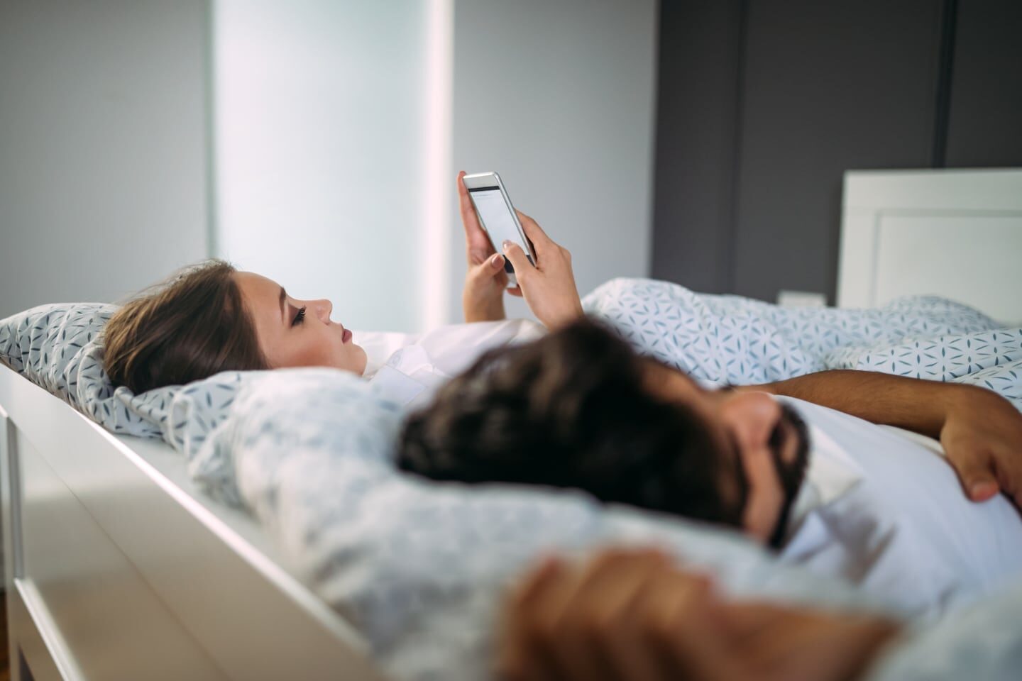 Pareja en la cama, uno revisando el teléfono y el otro durmiendo
